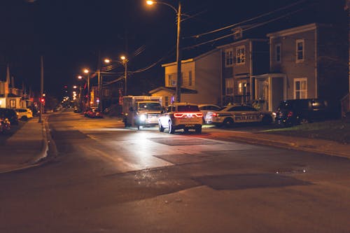Cars with glowing lights parked on asphalt road near police car placed near residential building in small town in evening