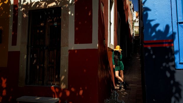 A woman in a green dress and yellow hat stands in a sunlit alleyway in Guanajuato, Mexico.