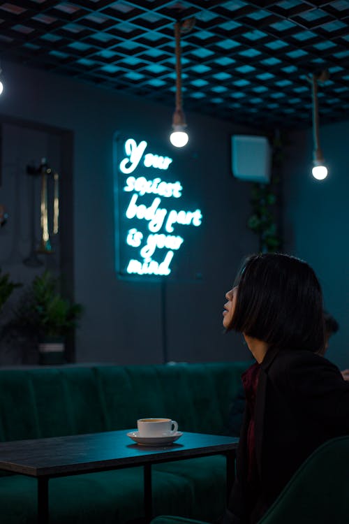 Side view of serious young female sitting at table with cup of hot drink in cafe with bright luminous inscription on wall