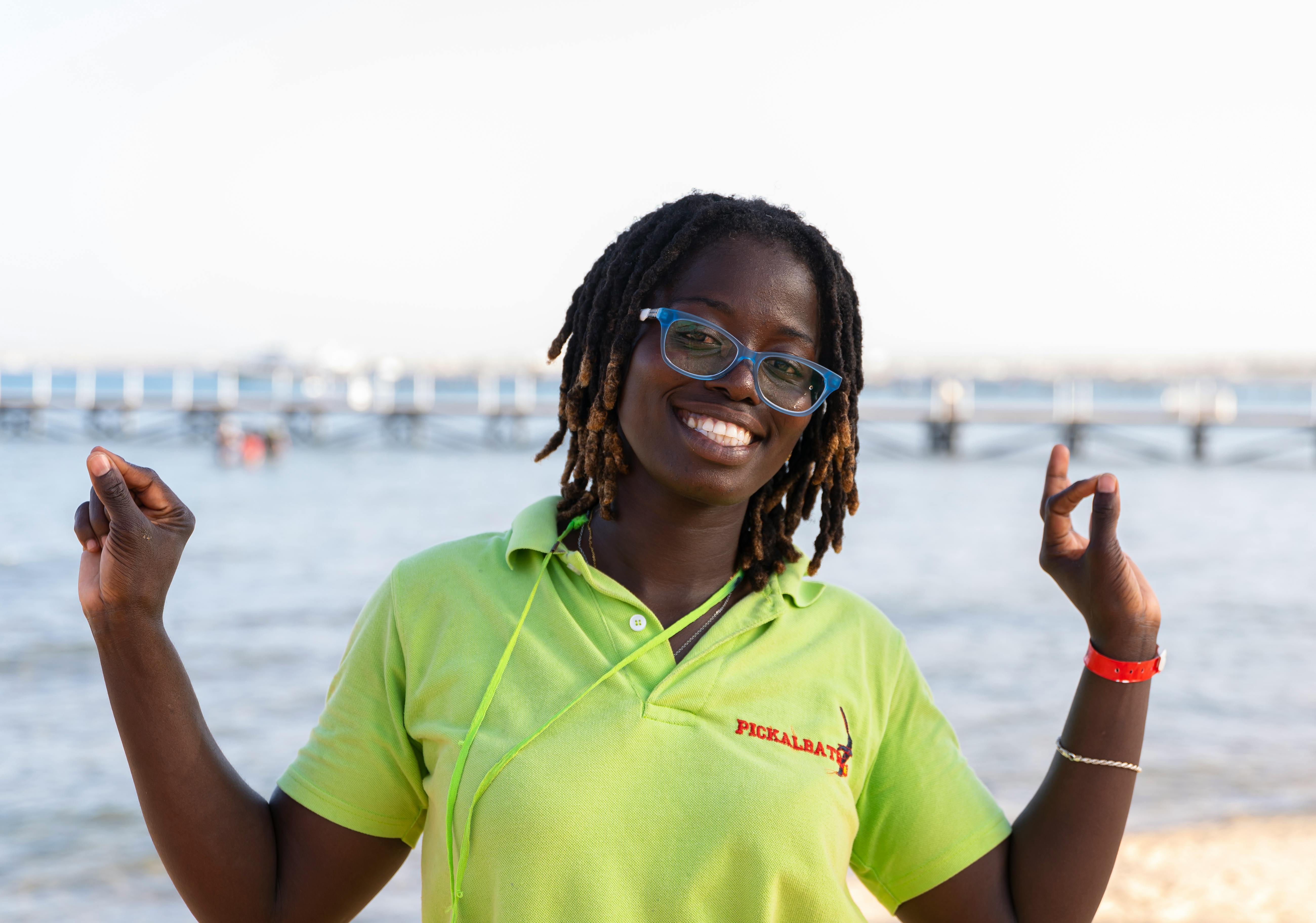 Woman smiling, raising arms, wearing glasses and green shirt, beach background.
