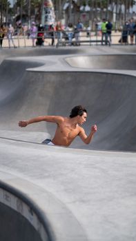 Young shirtless male skateboarder accelerating to jump on ramp in skater park while people standing and watching around