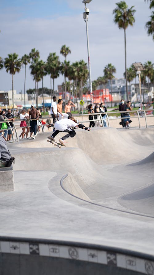 Free Anonymous person wearing elbow and knee laps performing stunt on ramp while people watching near fence with palms on background Stock Photo