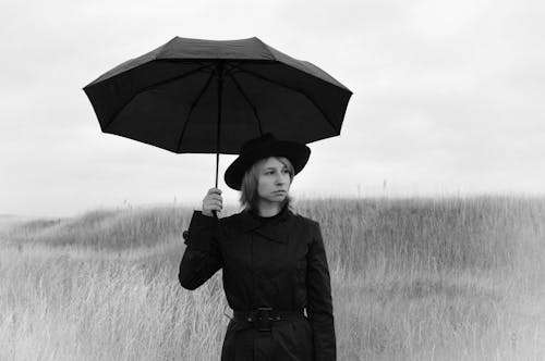 Black and white of serious pensive young female in coat and hat with umbrella looking away while standing on grassy field in gloomy rainy day