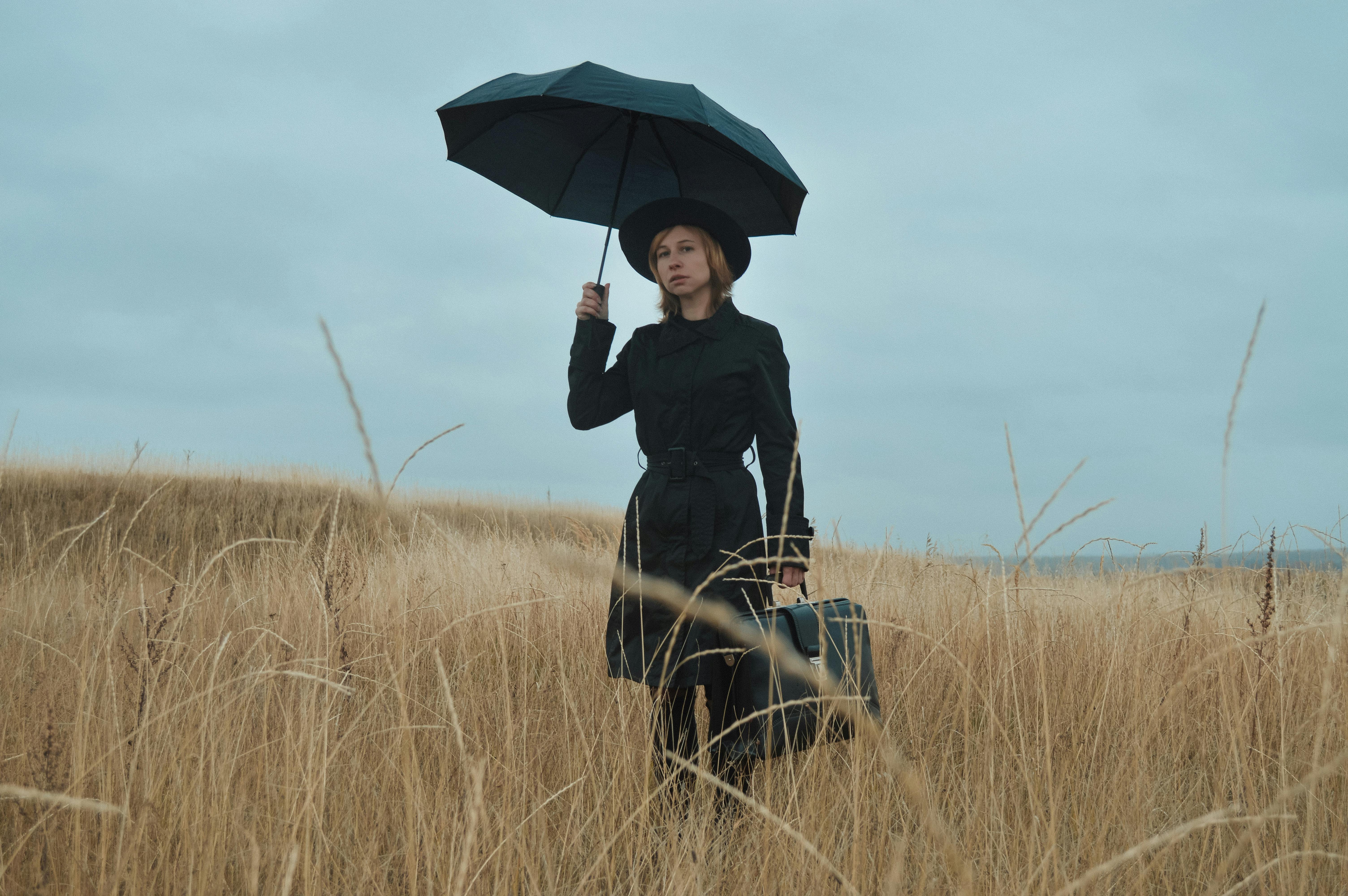 Serious young female in black coat and hat with umbrella and briefcase standing among dry grass in field and looking away in overcast weather