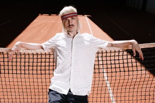 Vintage-inspired tennis player with a sweatband leaning on the net under sunlight on a court.