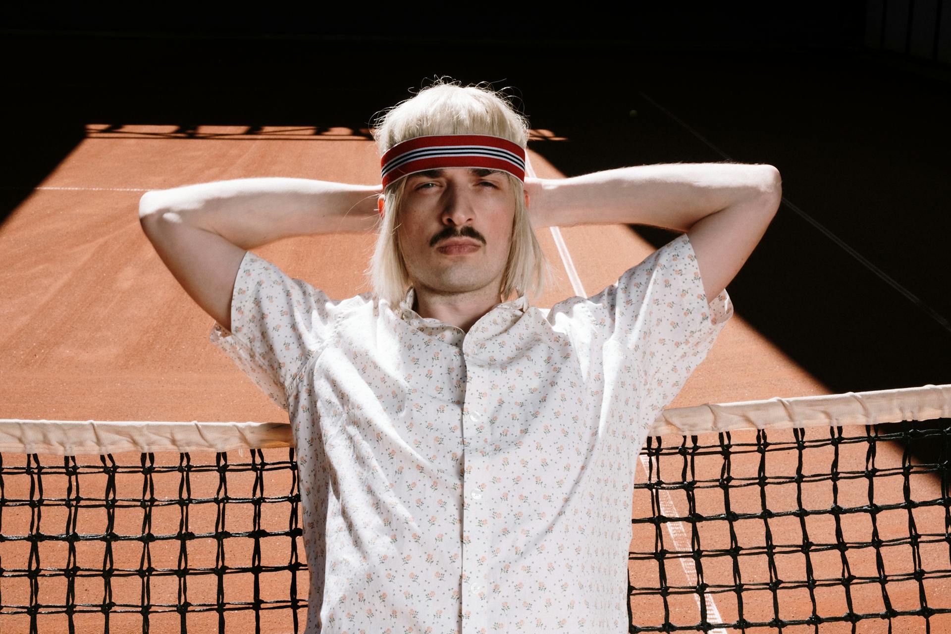 Caucasian man with mustache and headband leans on tennis net on sunlit court.