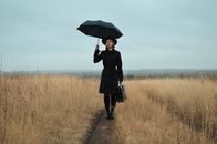 一位穿着黑色外套、戴着帽子的冷静年轻女性，手持雨伞和公文包，正全神贯注地站在干旱田野间的狭窄小路上，注视着镜头