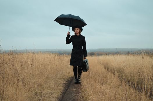 一位穿着黑色外套、戴着帽子的冷静年轻女性，手持雨伞和公文包，正全神贯注地站在干旱田野间的狭窄小路上，注视着镜头