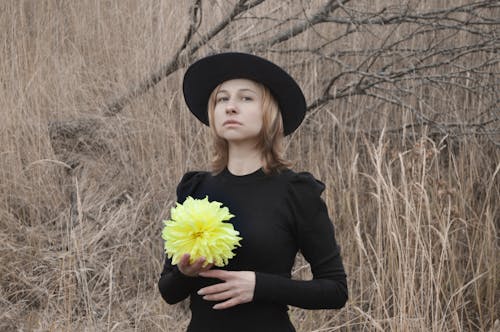 Tranquil elegant young female with blond hair in classic dress and hat standing near dry tree with flower in hand and looking at camera on gloomy autumn day