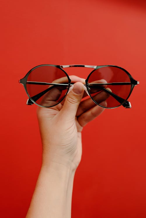 Crop anonymous person demonstrating stylish sunglasses in metal rim against red background in studio