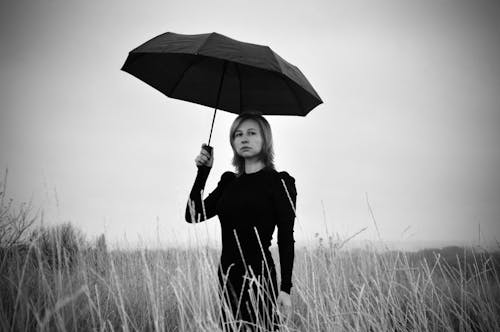 Black and white of unhappy young lady in elegant dress with umbrella in hand standing in rural terrain and looking at camera against overcast sky