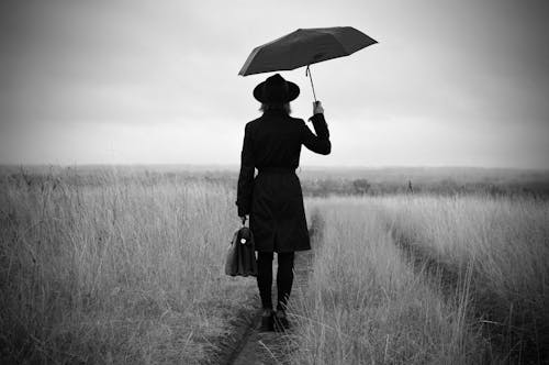 Back view of black and white anonymous lady in stylish coat and hat standing alone with umbrella and briefcast against cloudy sky in countryside