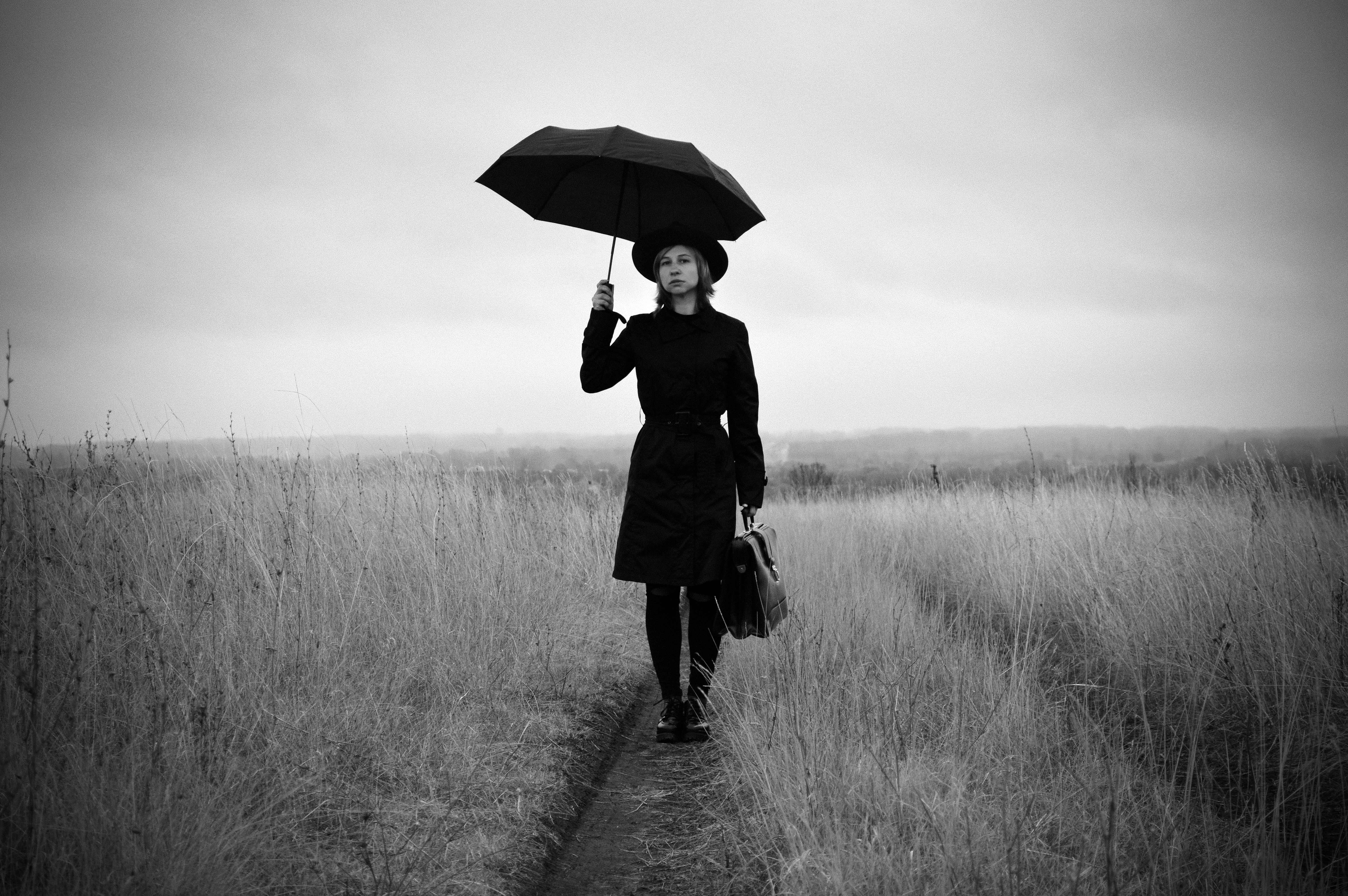 一位身着时尚外套和帽子、手持雨伞与公文包的年轻女士，平静地站在干燥的草地上，她全身黑白相间的装束映衬着乡村多云的天空