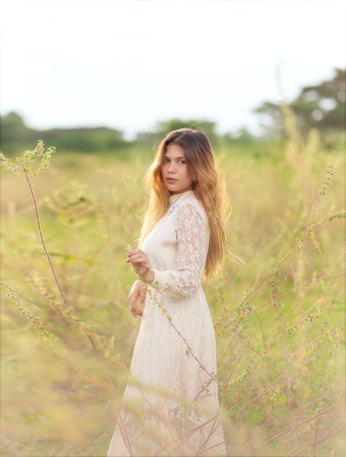 Woman in White Long Dress Standing on Green Field while Seriously Looking at the Camera
