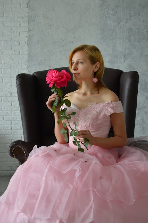 Content young bride in elegant pink dress sitting in comfortable armchair and enjoying smell of fresh rose