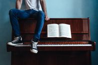 Crop unrecognizable male in trendy gumshoes and jeans sitting on piano with sheet music in classroom