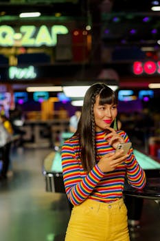 Asian woman in a vibrant striped outfit uses smartphone at an indoor amusement center, blending fashion and technology.