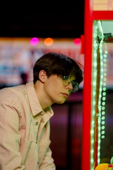 Portrait of a young man with eyeglasses focused on an arcade game in a vibrant setting.