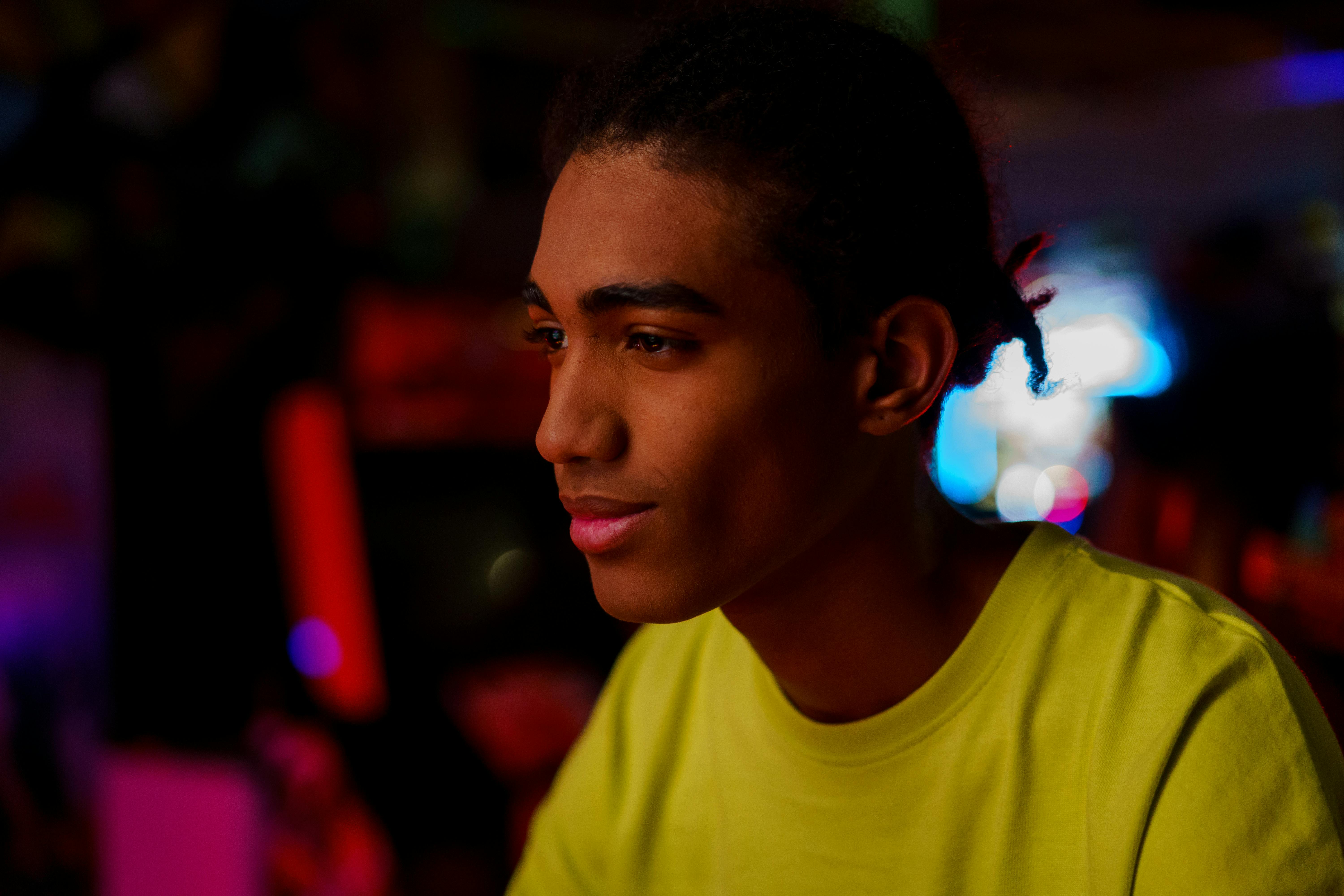A young man wearing a yellow t-shirt smiling under vibrant neon lighting indoors.