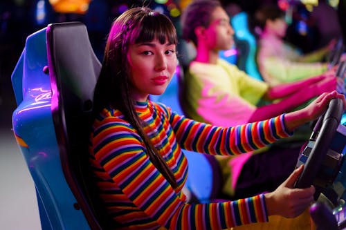 A Woman in Striped Long Sleeves Sitting on a Car Arcade Machine while Seriously Looking at the Camera