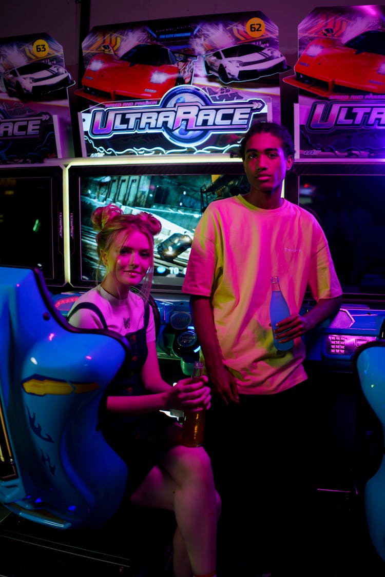 A Young Man And Woman Near An Arcade Machine Holding A Bottle Of Soda While Looking At The Camera