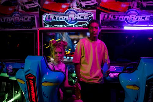 Man Standing Beside a Woman Sitting on a Car Arcade Machine Holding a Bottle of Soda while Looking at the Camera