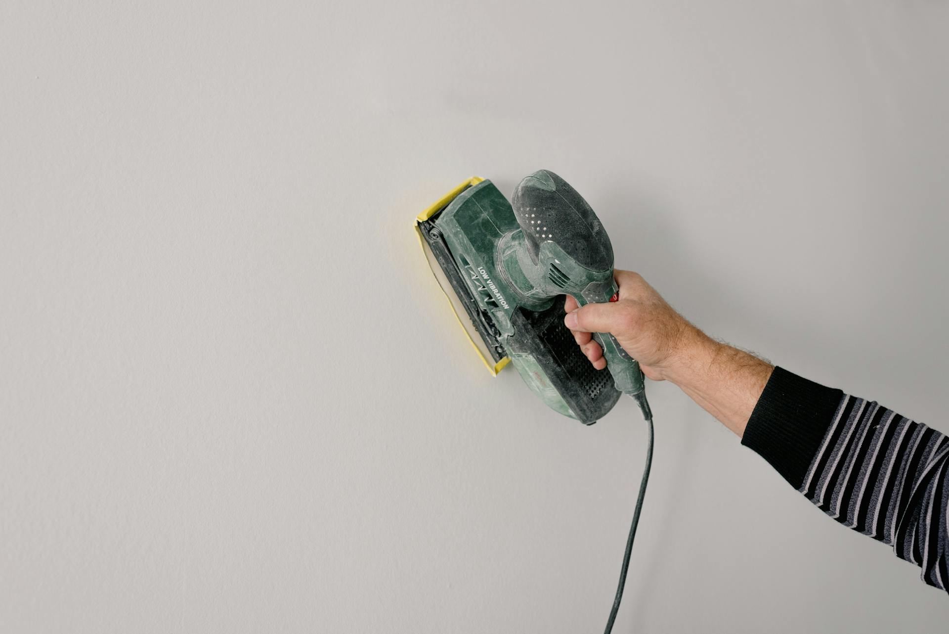 A person uses an electric sander to smooth a wall indoors, showcasing home renovation skills.