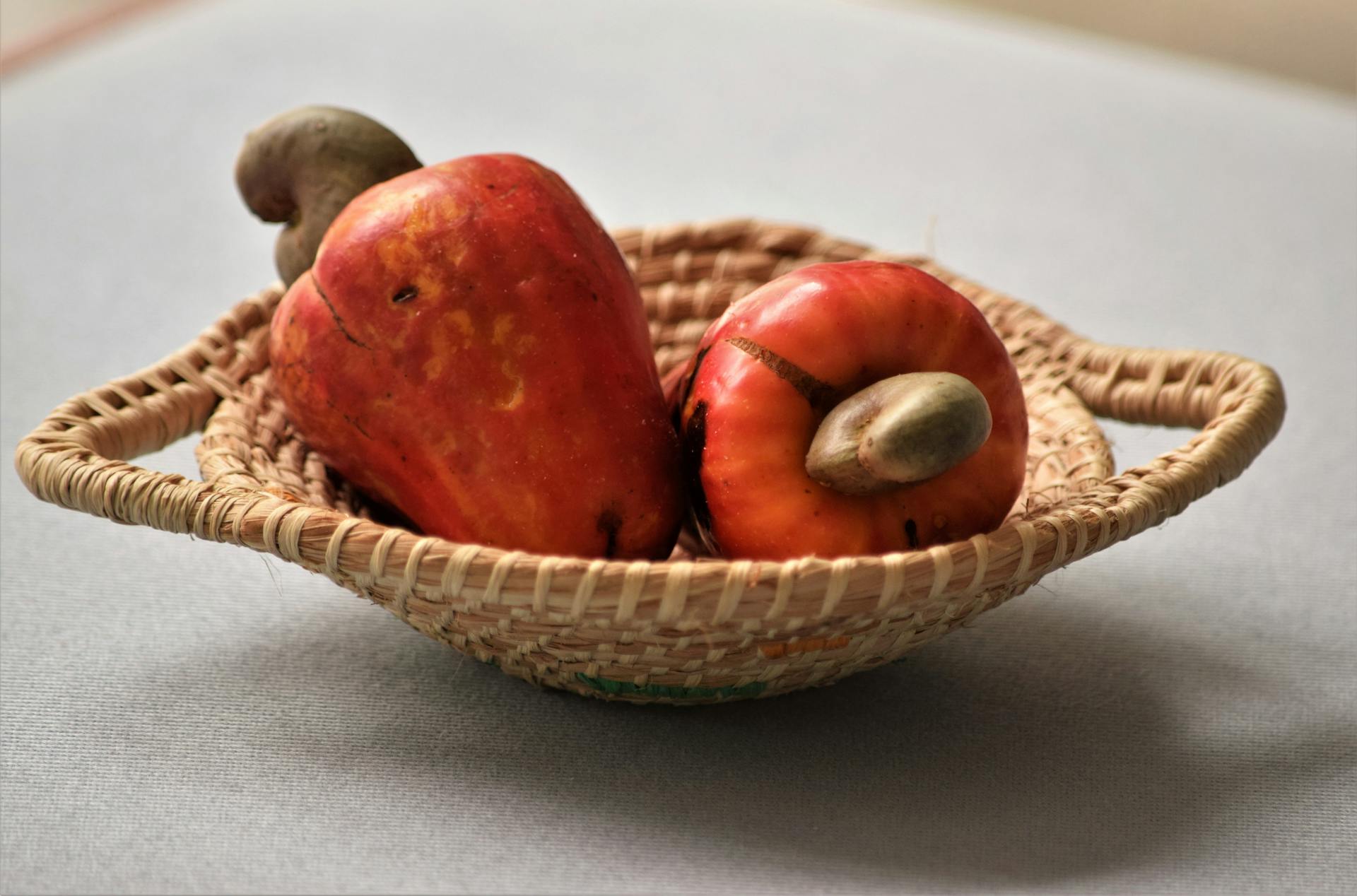 Close-up of ripe cashew apples in a woven basket on a white surface, perfect for food and natural settings.