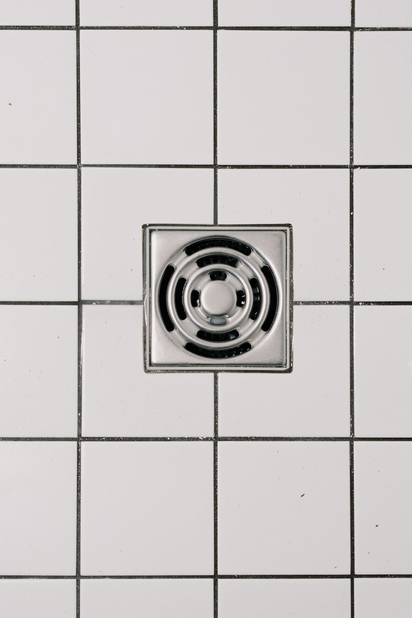 Close-up of a metal floor drain installed within square white floor tiles with dark grout lines.