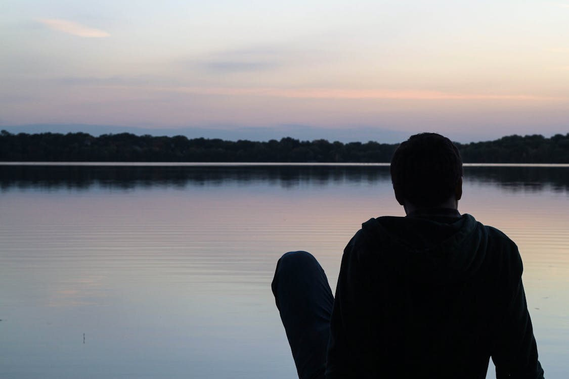 Фотография силуэта человека в толстовке, сидящего перед большим водоемом во время заката