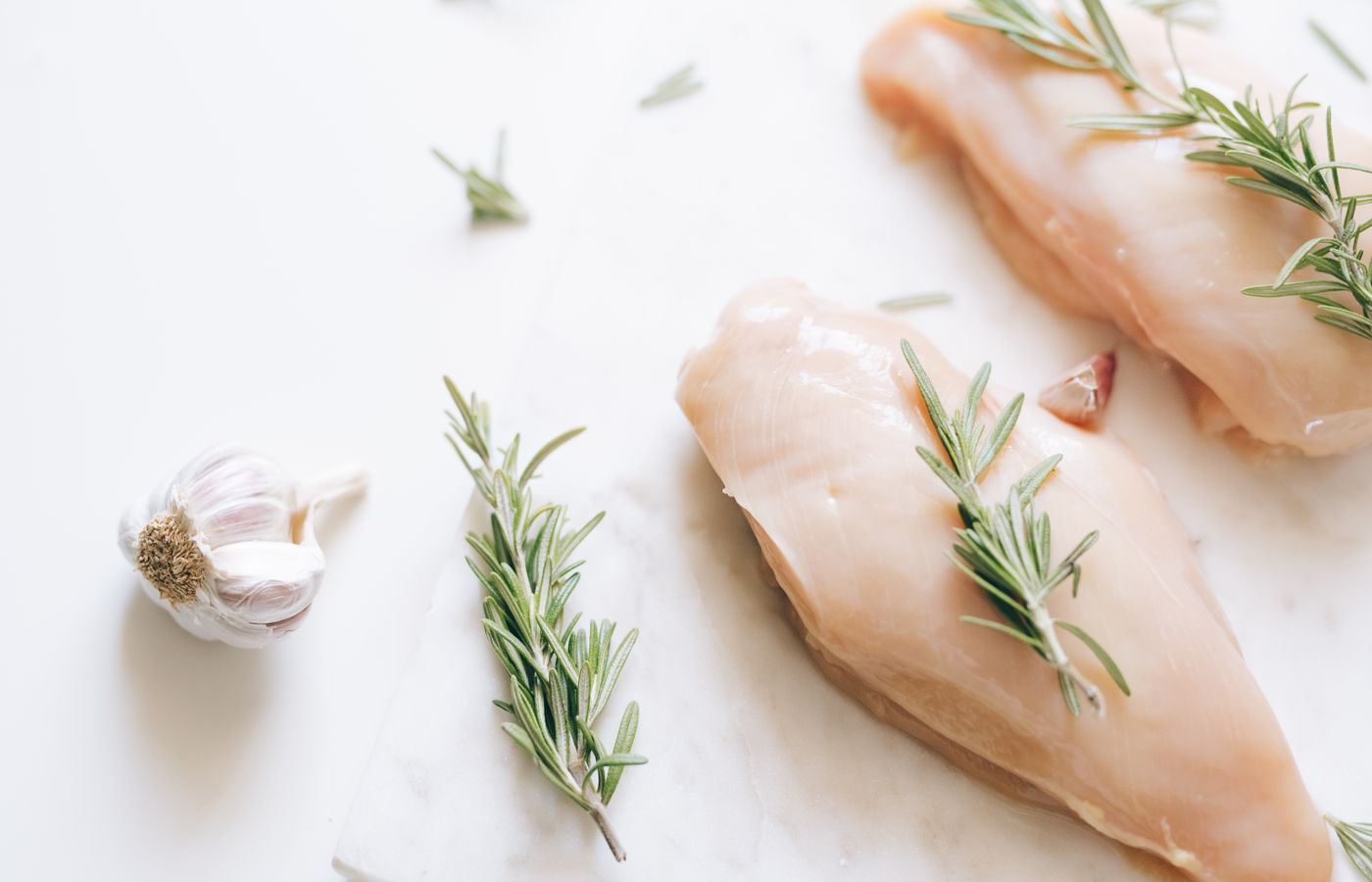 Raw chicken breasts garnished with rosemary and garlic on a white surface.