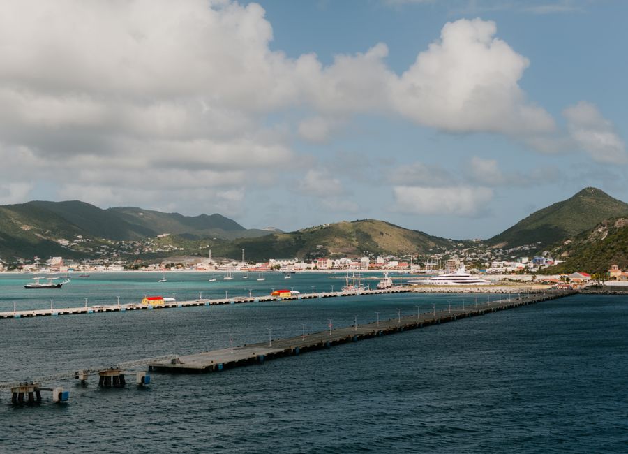 Tranquil harbor scene with piers, mountains, and vibrant town of St. Maarten under a bright sky.