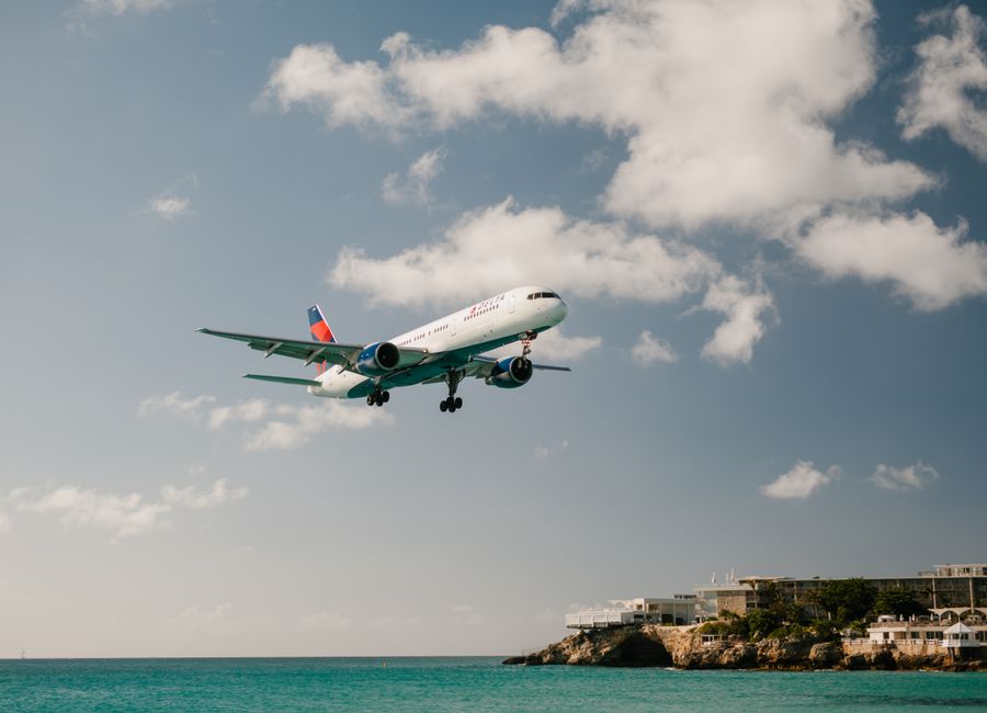 Commercial aircraft landing above ocean with clear blue skies, near beach resort.