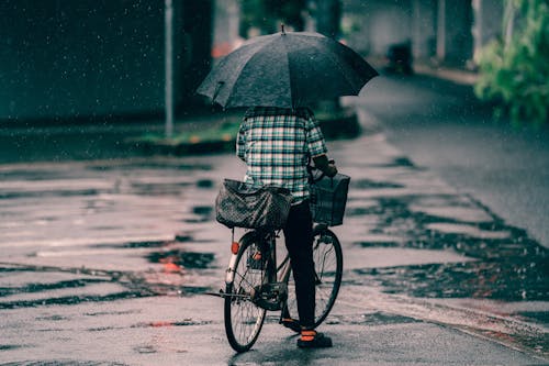 Person in Plaid Shirt and Black Pants Holding an Umbrella While Riding a Bicycle