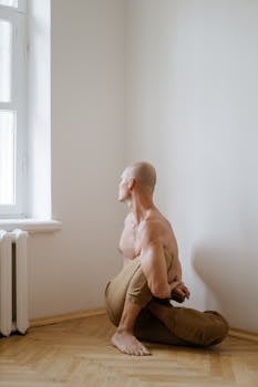 Bald man in a seated yoga pose gazing out a window, promoting peaceful wellness.