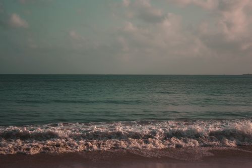 Free Ocean waves crash gently on a sandy beach under a cloudy sky at sunset, creating a tranquil seascape. Stock Photo