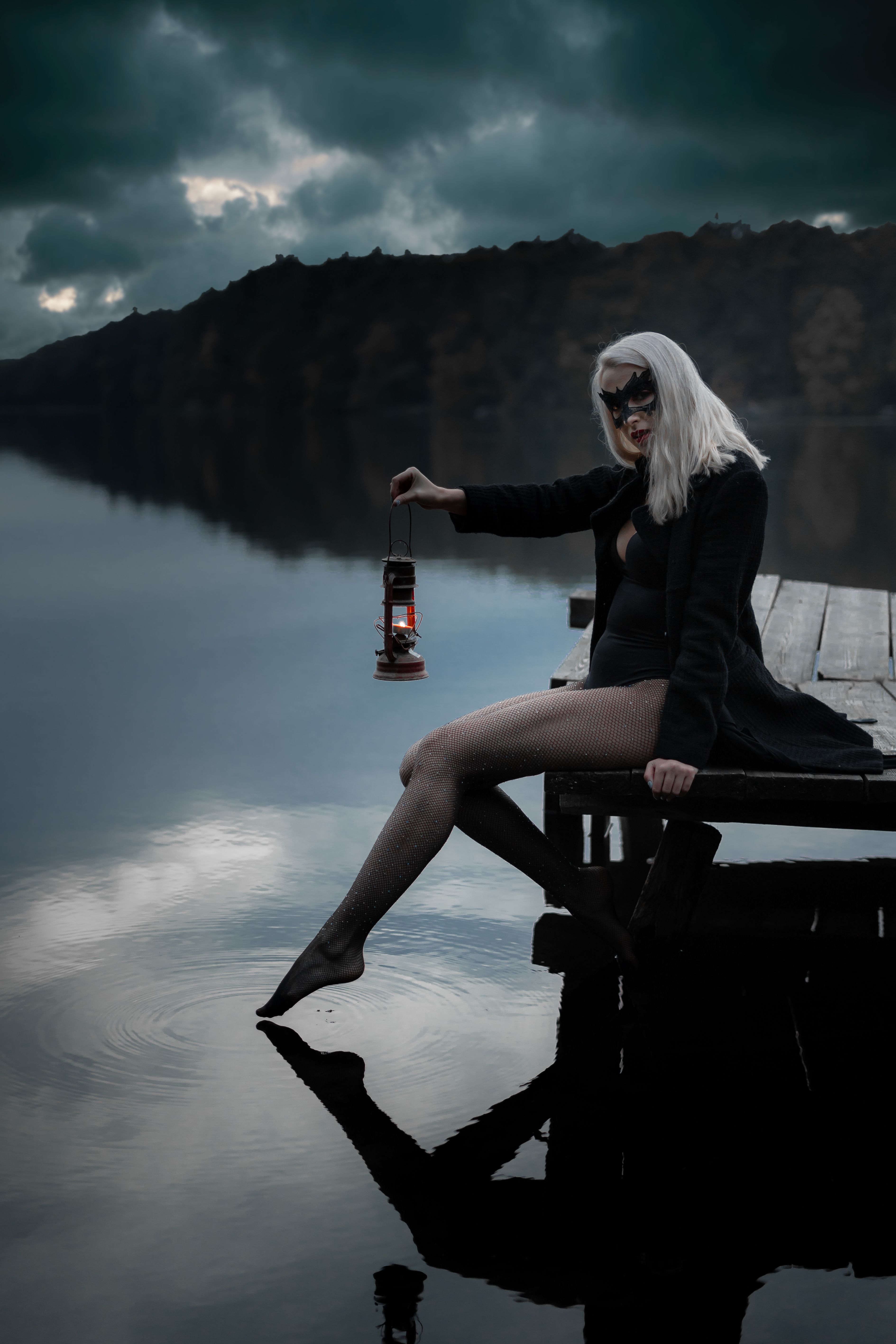 Side view of blond female in trendy apparel and decorative mask reflecting in river while sitting on wooden dock and looking at camera at dusk