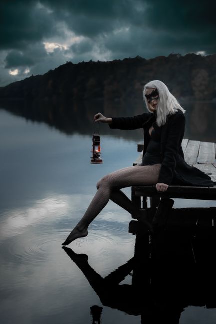 Side view of blond female in trendy apparel and decorative mask reflecting in river while sitting on wooden dock and looking at camera at dusk