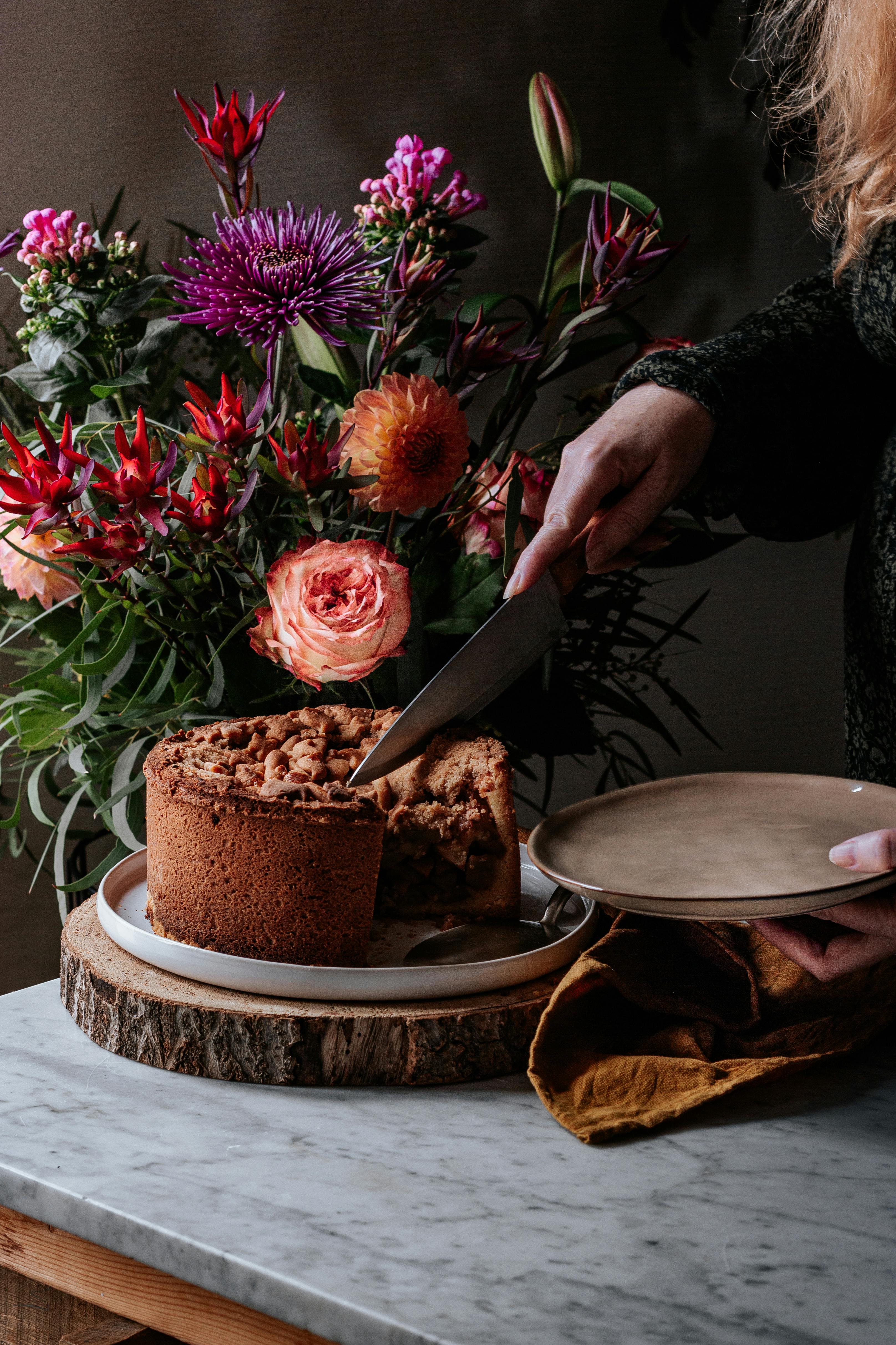 Person slicing a cake on a wooden board with a bouquet of flowers in the background.