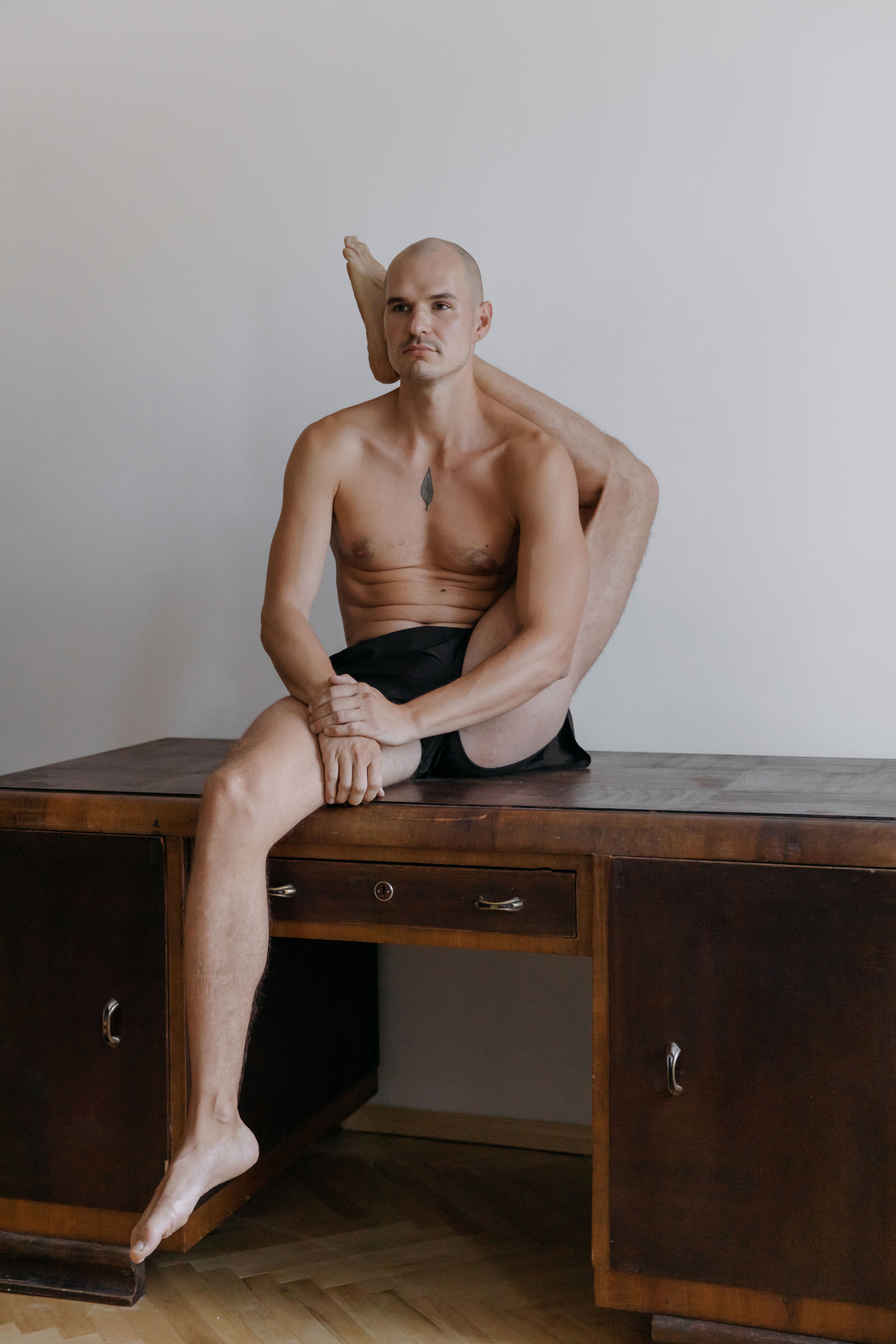 A serious shirtless man with a flexible posture sits comfortably on a wooden desk.