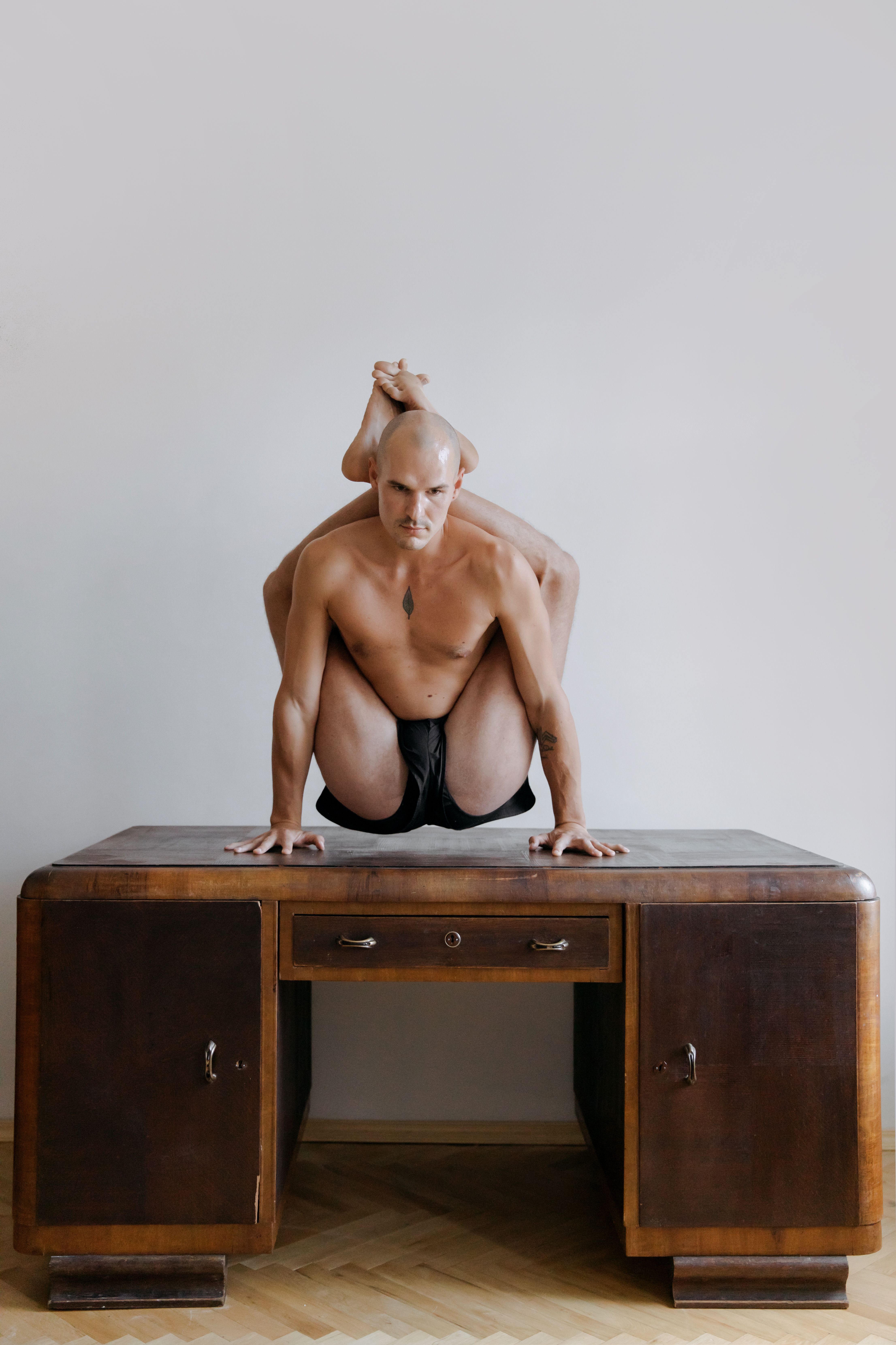 Flexible bald man balancing on a wooden desk demonstrating advanced yoga pose indoors.