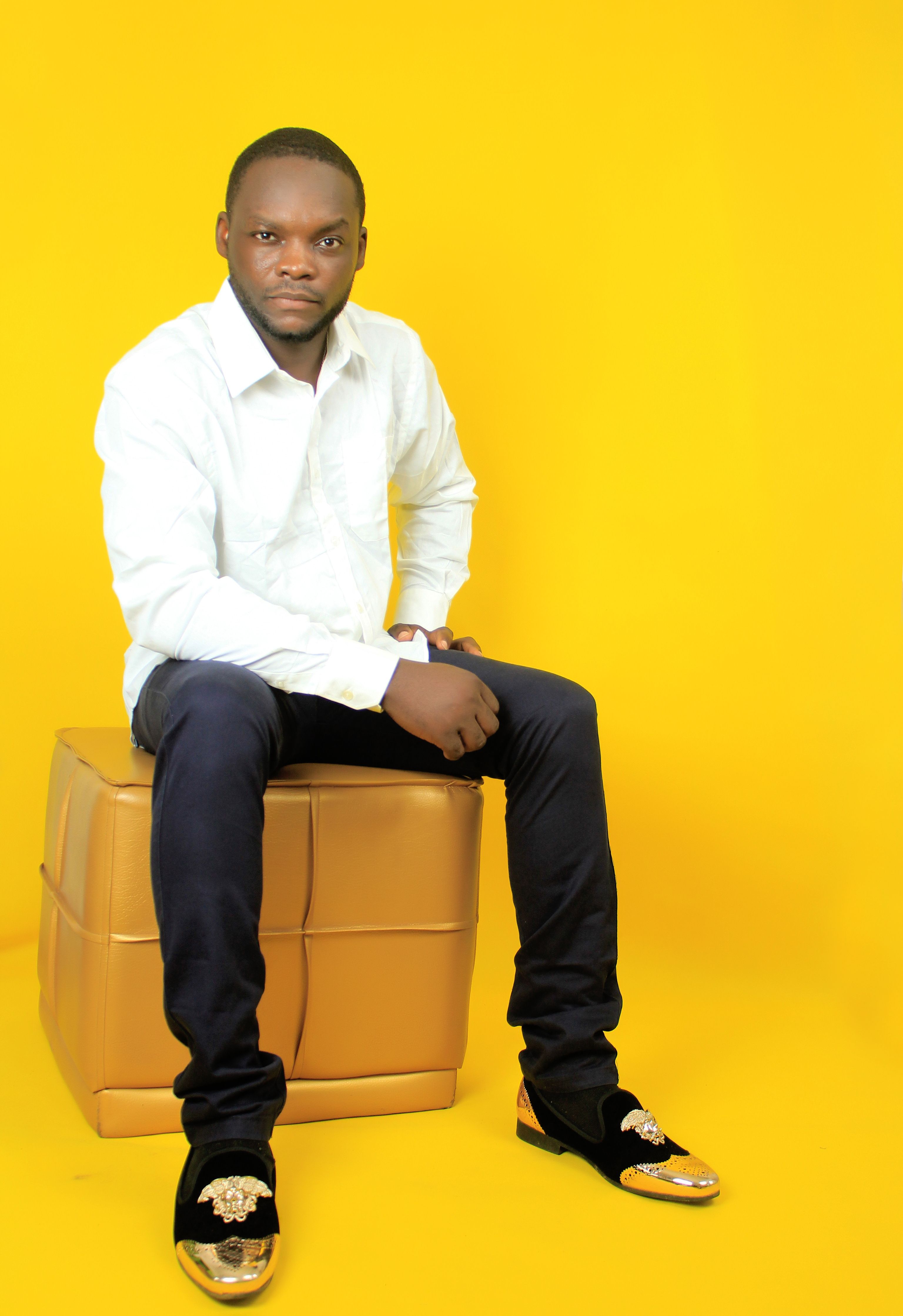 Stylish man in white shirt sitting on a tan ottoman against a vibrant yellow backdrop.