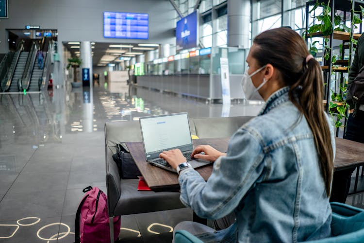 Focused Woman In Mask Browsing Laptop In Airport