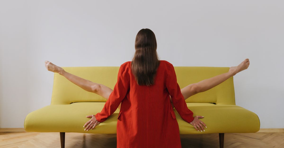 Femme en blazer rouge assise de manière créative sur un canapé jaune avec un mur blanc en arrière-plan.