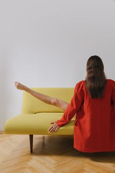一位身着红色西装的女士在室内的黄色沙发上舒展身体，展现了她的灵活与风格