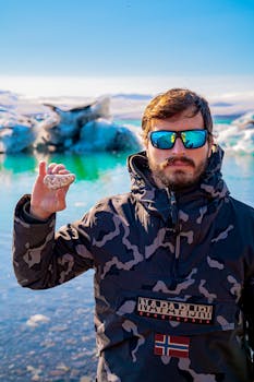 A bearded man in sunglasses and camouflage jacket holding a rock near icy waters.