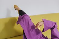 一位时尚女性穿着紫色外套和黑色紧身裤，正躺在一张现代黄色沙发上，显得既时尚又放松