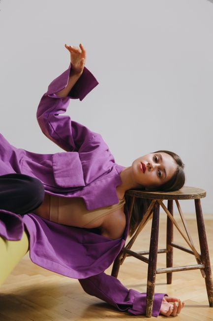 Artistic portrayal of a woman in a vibrant purple coat reclined on a wooden stool.