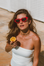 From above of calm female in red sunglasses looking at camera while standing on tiled floor with half of orange near white fence in sunlight