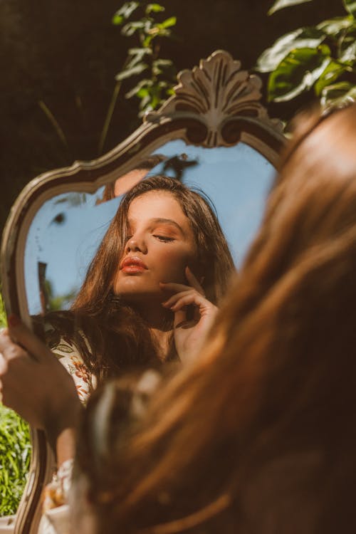 Attractive dreamy female with hand on cheek reflecting in mirror and looking away while sitting in nature with green foliage in summer